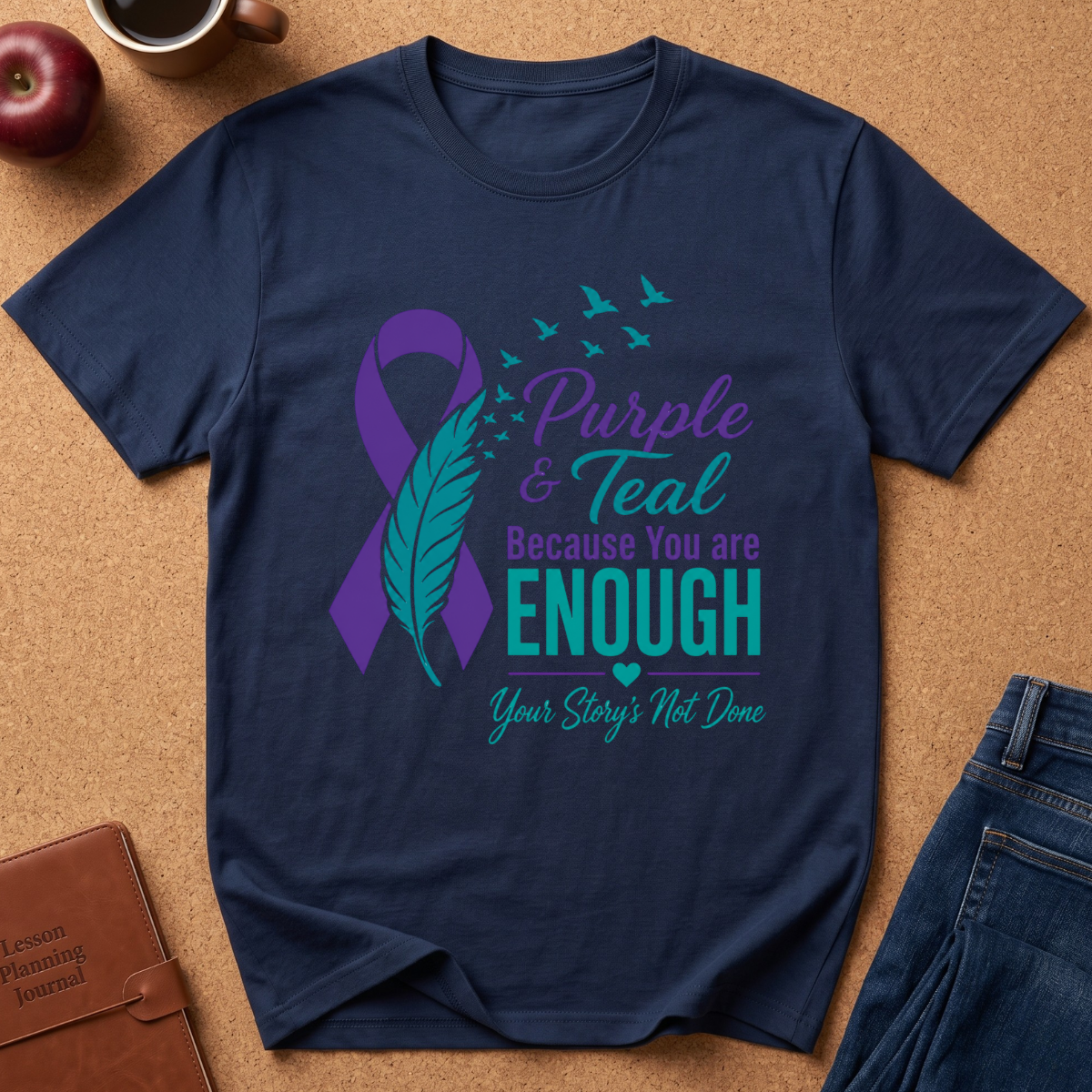 A navy blue short-sleeve t-shirt with a purple and teal awareness graphic on the front, laid flat on a brown cork background with a coffee cup, apple, and jeans nearby.