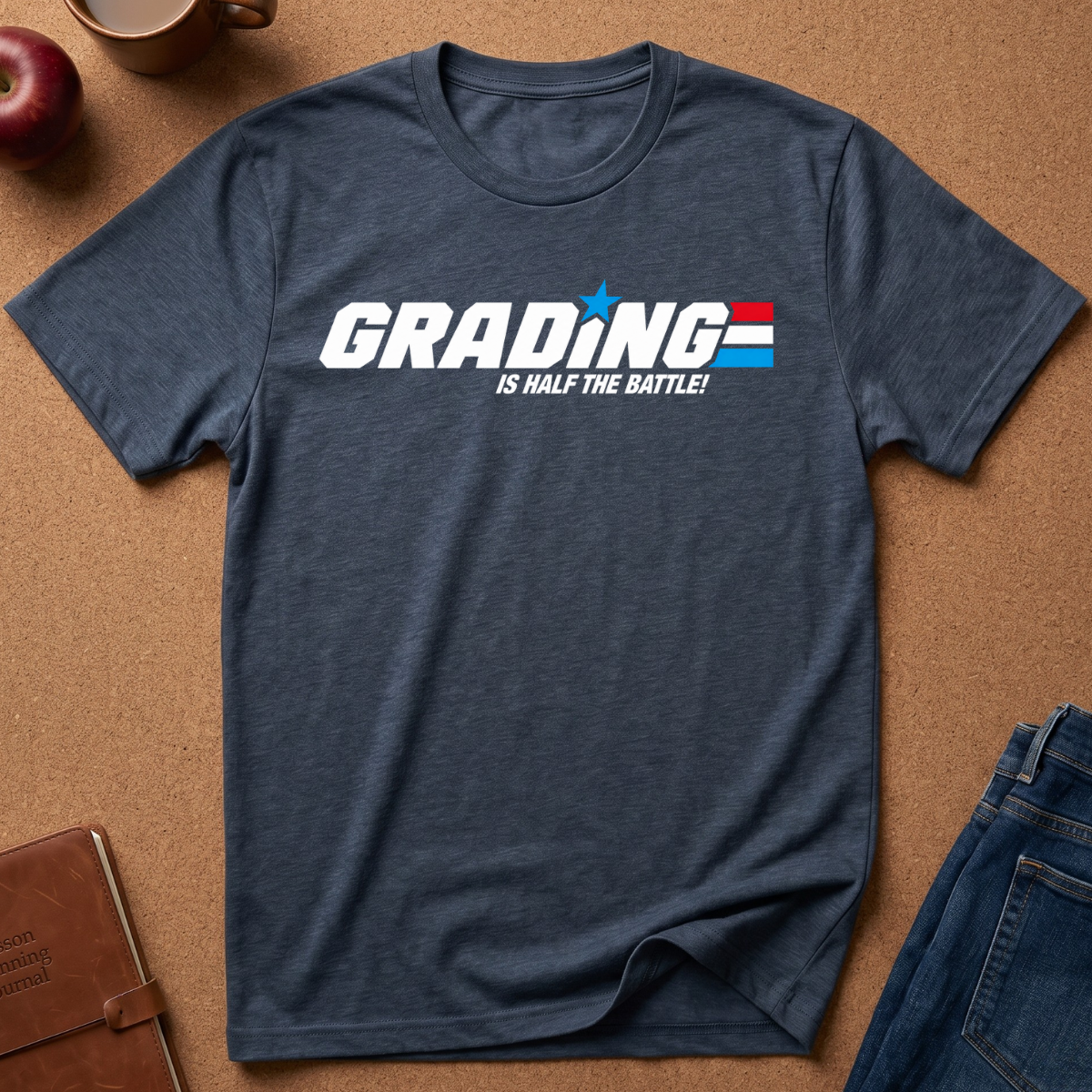 A navy blue heather graphic t-shirt laid flat on a brown cork background, styled with a notebook, apple, coffee cup, and jeans. The shirt features a white 'GRADING' text graphic with a blue star and red/white/blue flag design, and the phrase 'IS HALF THE BATTLE!' underneath.