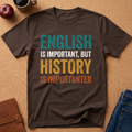 A dark brown graphic t-shirt laid flat on a cork background with lifestyle props including an apple, a coffee mug, a journal, and blue jeans. The shirt features a humorous text design that reads 'ENGLISH IS IMPORTANT, BUT HISTORY IS IMPORTANTER' in teal, white, yellow, and orange distressed lettering.