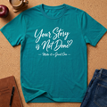 A teal short-sleeve graphic T-shirt laid flat on a cork surface, with white script text reading 'Your Story is Not Done? Make it a Great One.' Surrounding props include a coffee cup, apple, journal, pen, and jeans.