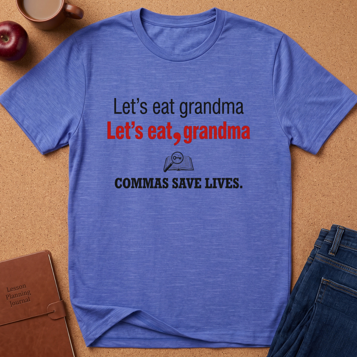 A heathered blue graphic t-shirt laid flat on a cork background, surrounded by lifestyle props including an apple, a coffee mug, a journal, and jeans. The shirt features the text 'Let's eat grandma Let's eat, grandma COMMAS SAVE LIVES.