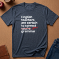 A heather navy blue graphic t-shirt laid flat on a brown cork background. The shirt features a white printed text design that reads 'English teachers are certain to correct you're grammar' with 'you're' crossed out in red script and 'your' written in red cursive.
