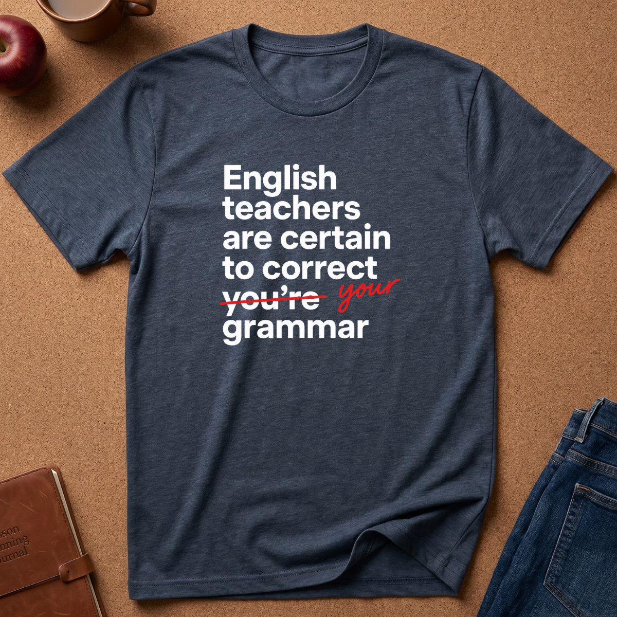 A heather navy blue graphic t-shirt laid flat on a brown cork background. The shirt features a white printed text design that reads 'English teachers are certain to correct you're grammar' with 'you're' crossed out in red script and 'your' written in red cursive.