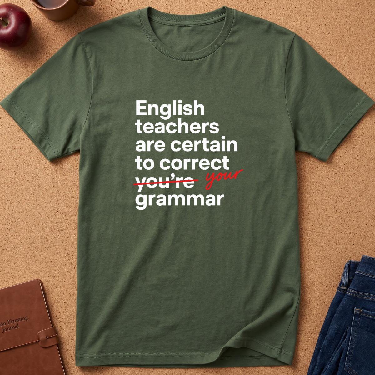 An olive green short-sleeve t-shirt with a white and red printed text graphic reading 'English teachers are certain to correct you're your grammar', laid flat on a cork background with a book, apple, and jeans as props.