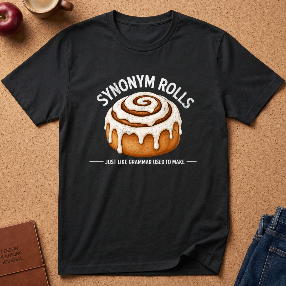 A black short-sleeve graphic T-shirt laid flat on a cork background, featuring a cinnamon roll illustration with the text 'SYNONYM ROLLS' above and 'JUST LIKE GRAMMAR USED TO MAKE' below. Styled flat-lay props include an apple, mug, journal, and jeans around the shirt.