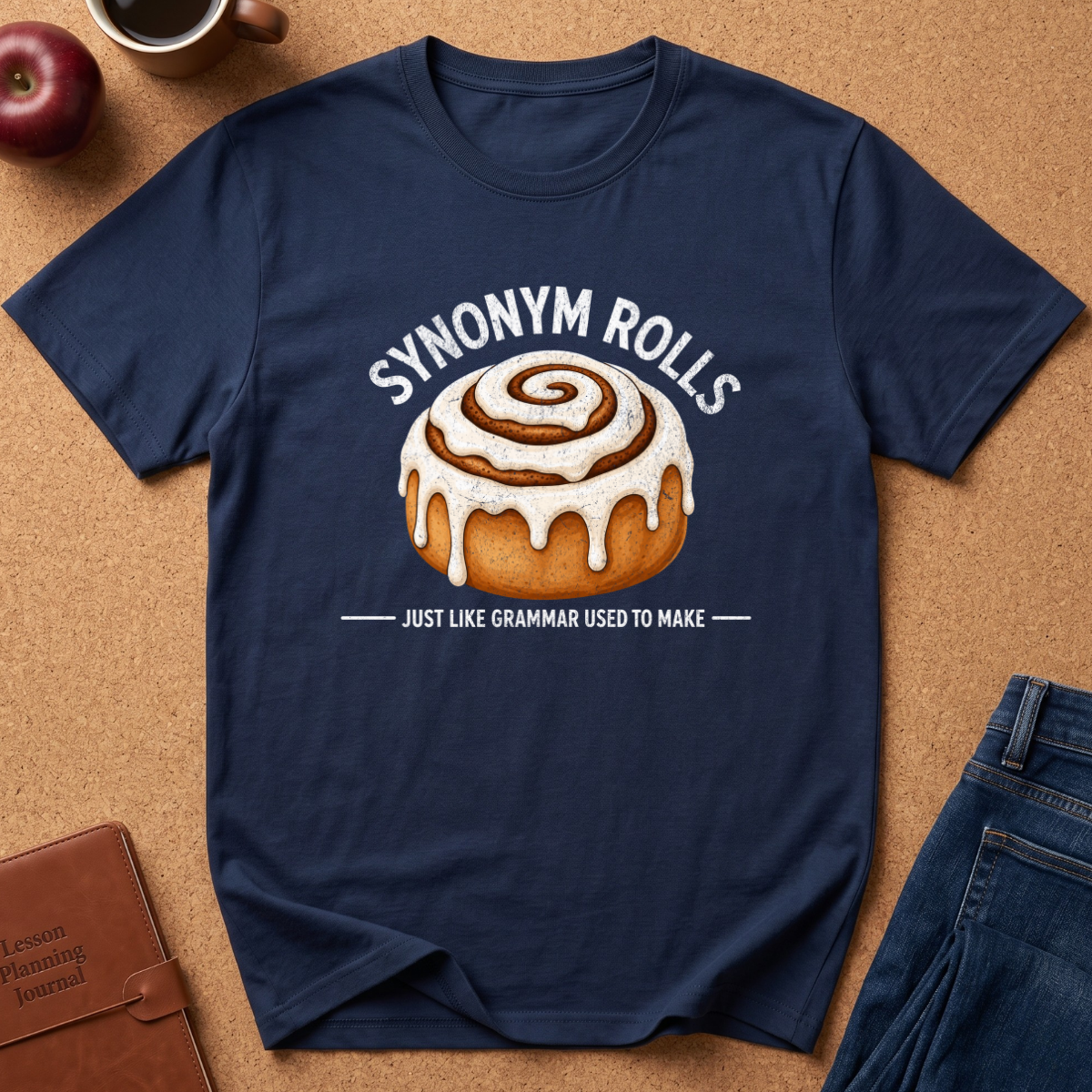 A navy blue graphic t-shirt laid flat on a brown cork background, featuring a cinnamon roll illustration and the text 'SYNONYM ROLLS' and 'JUST LIKE GRAMMAR USED TO MAKE'. Surrounding props include an apple, a coffee cup, a brown journal, and blue jeans.