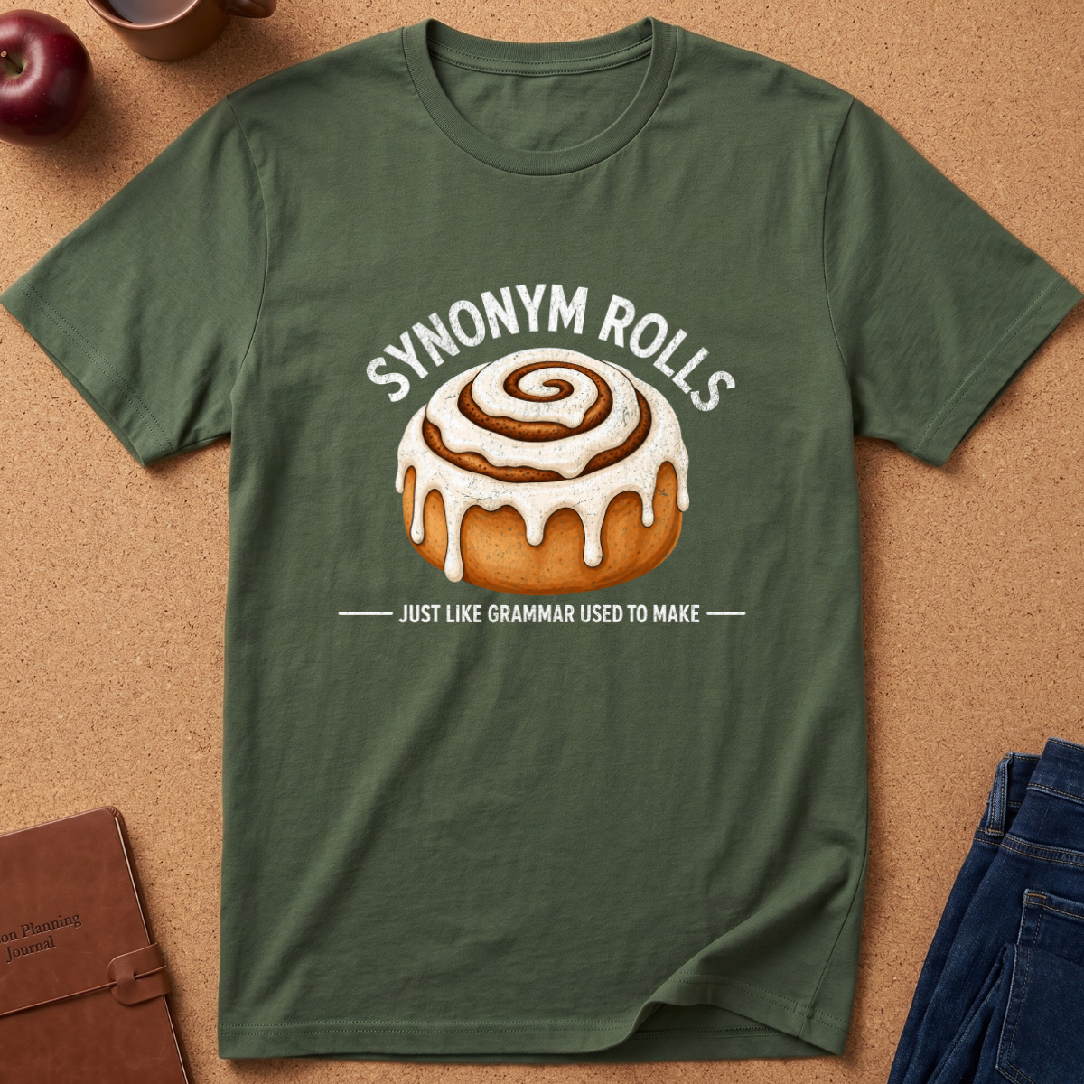 An olive green short-sleeve t-shirt laid flat on a cork background, featuring a graphic of a cinnamon roll with the text 'SYNONYM ROLLS' above and 'JUST LIKE GRAMMAR USED TO MAKE' below.