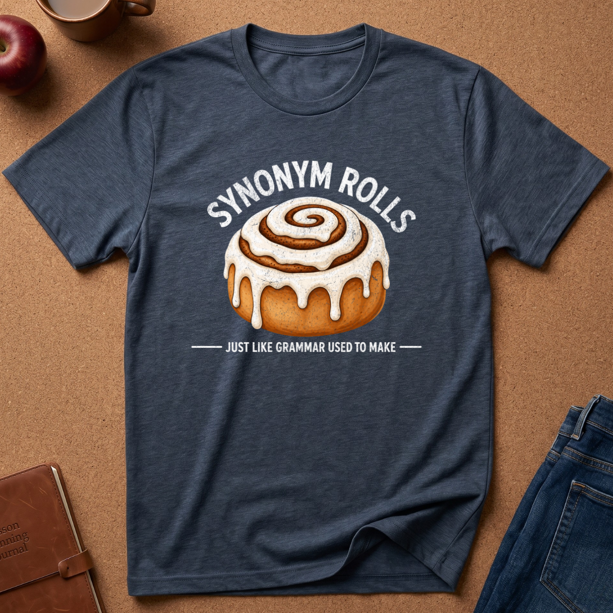 A heathered navy blue t-shirt laid flat on a brown cork background, featuring a graphic of a cinnamon roll with the text 'SYNONYM ROLLS' and 'JUST LIKE GRAMMAR USED TO MAKE'.