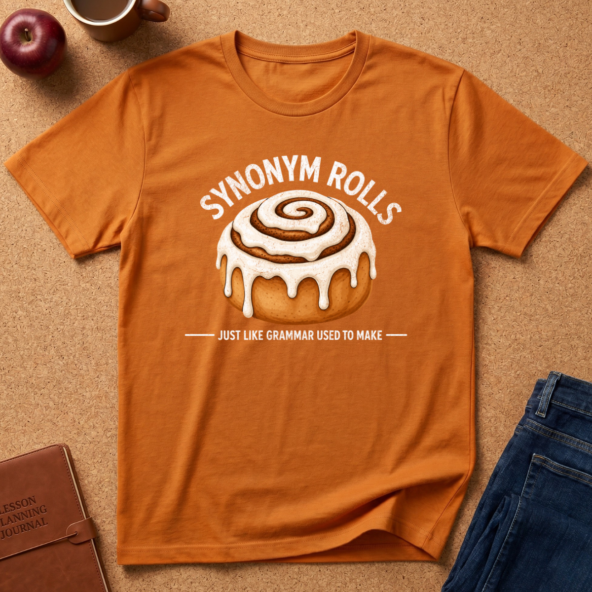 An orange crewneck t-shirt with a white graphic of a cinnamon roll and the text 'SYNONYM ROLLS' and 'JUST LIKE GRAMMAR USED TO MAKE', laid flat on a cork background with lifestyle props including an apple, a notebook, and jeans.