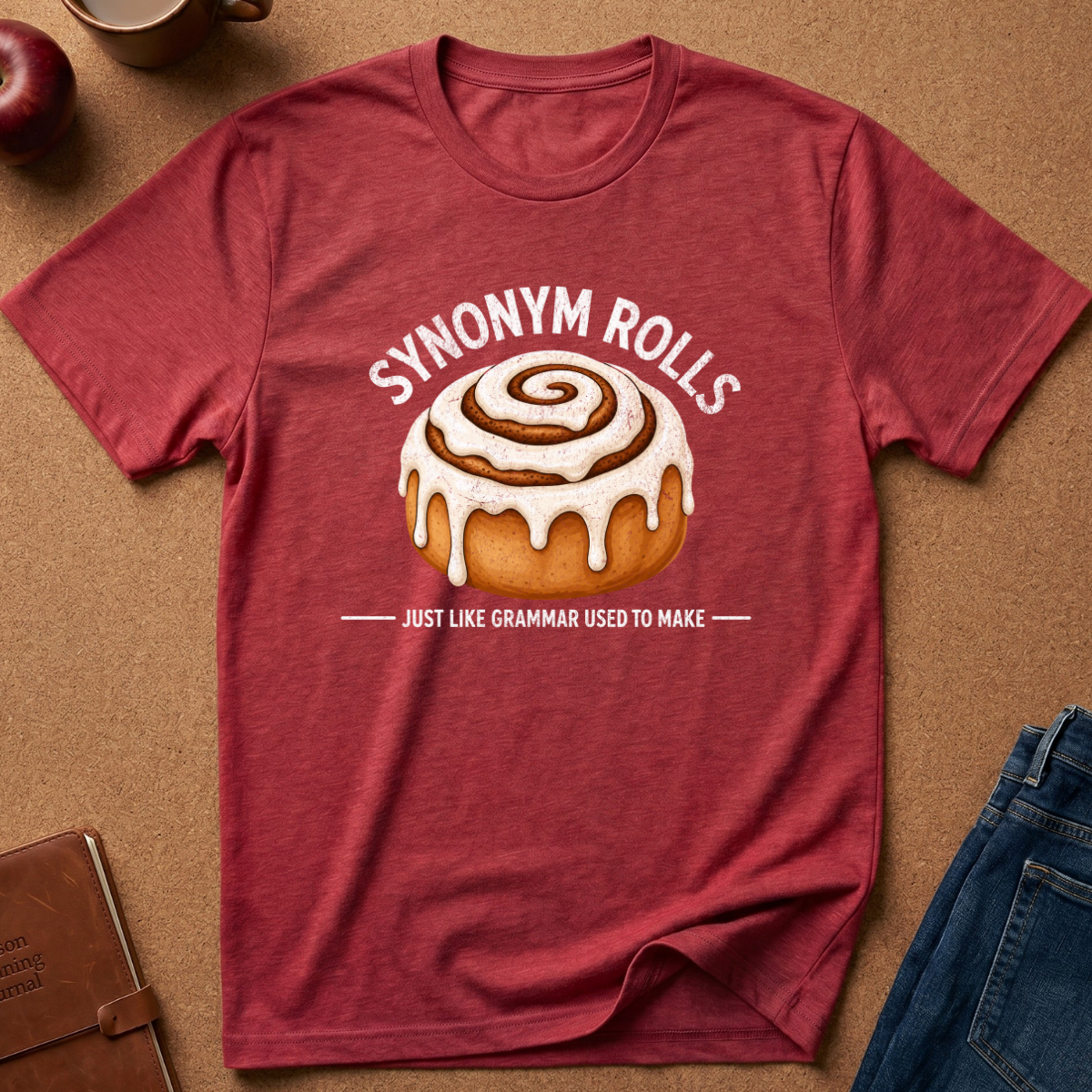 A heathered red graphic t-shirt laid flat on a brown cork background, featuring a cinnamon roll illustration and the text 'SYNONYM ROLLS' and 'JUST LIKE GRAMMAR USED TO MAKE'.