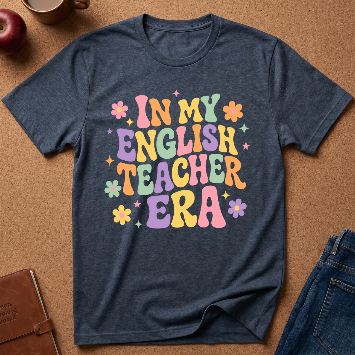 A navy blue heather t-shirt with a colorful retro-style graphic reading 'IN MY ENGLISH TEACHER ERA' in pastel letters, surrounded by small flowers and stars. The shirt is laid flat on a brown cork background with lifestyle props including an apple, a mug, a notebook, and jeans.
