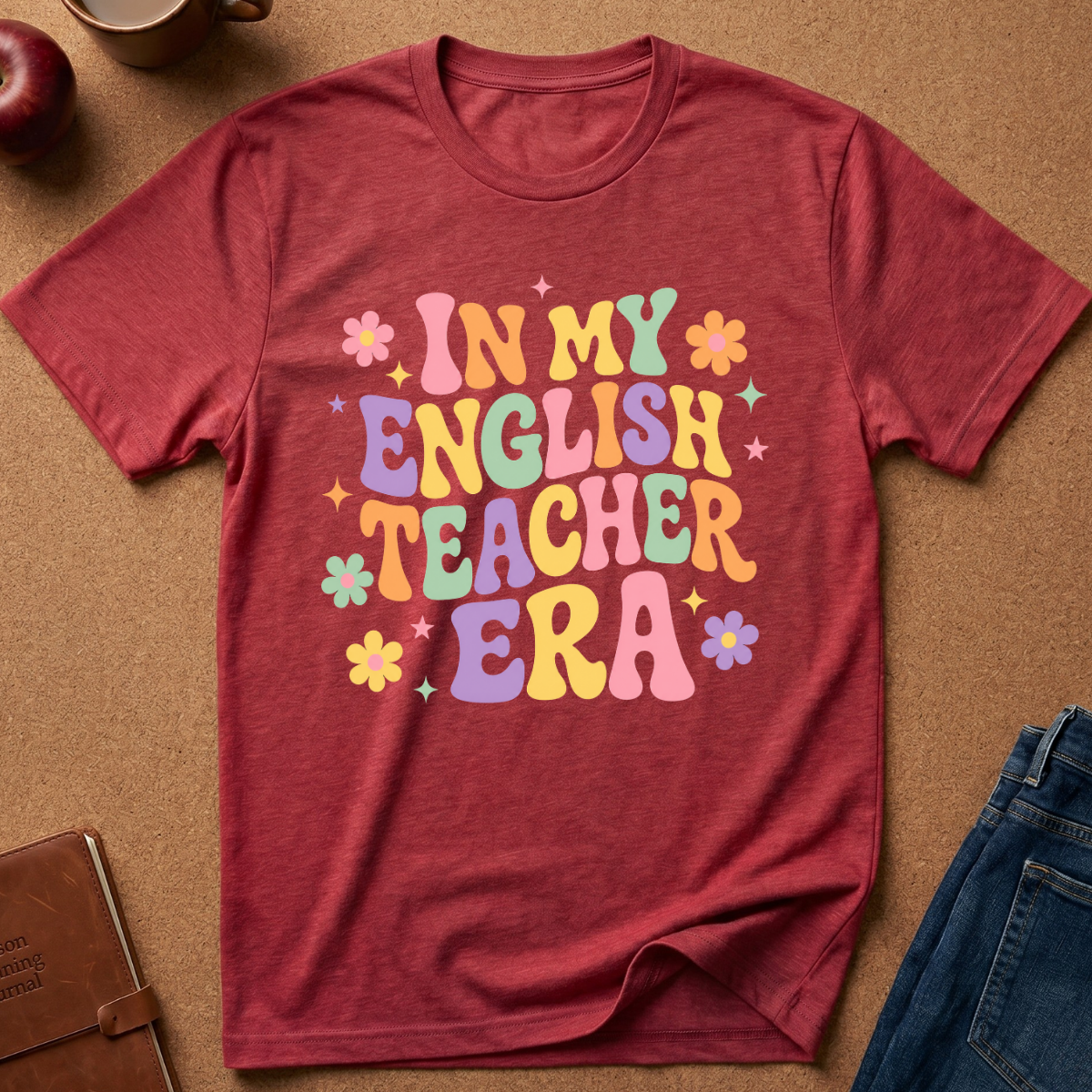 A heathered red short-sleeve t-shirt with a colorful retro-style graphic that reads 'IN MY ENGLISH TEACHER ERA' in pastel letters, surrounded by small flowers and stars. The shirt is laid flat on a brown cork background with lifestyle props including jeans, a notebook, a coffee mug, and an apple.