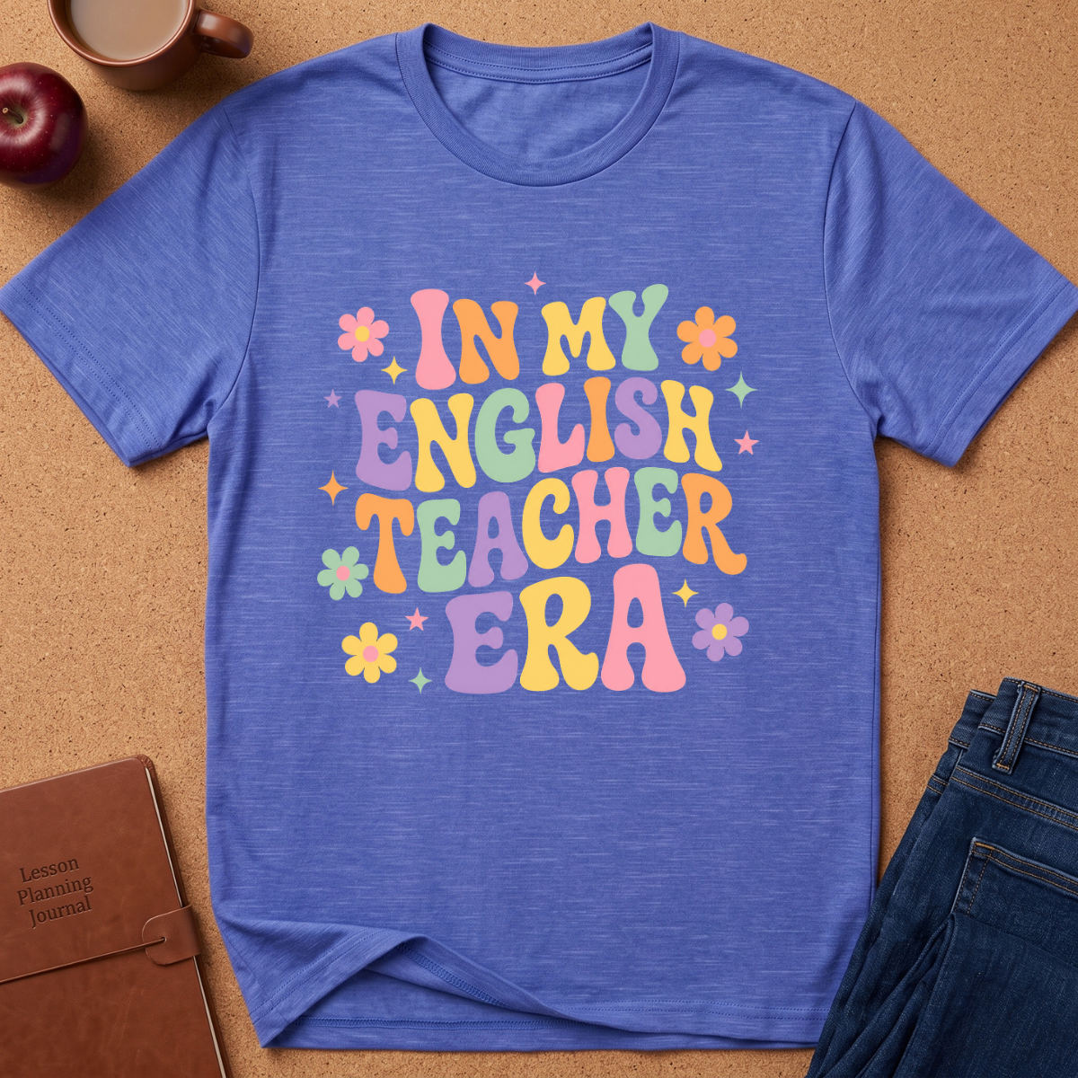 A heathered blue short-sleeve t-shirt with a colorful retro-style graphic reading 'IN MY ENGLISH TEACHER ERA' in pastel letters, surrounded by small flowers and stars. The shirt is laid flat on a cork background with a coffee mug, apple, notebook, and jeans partially visible around it.