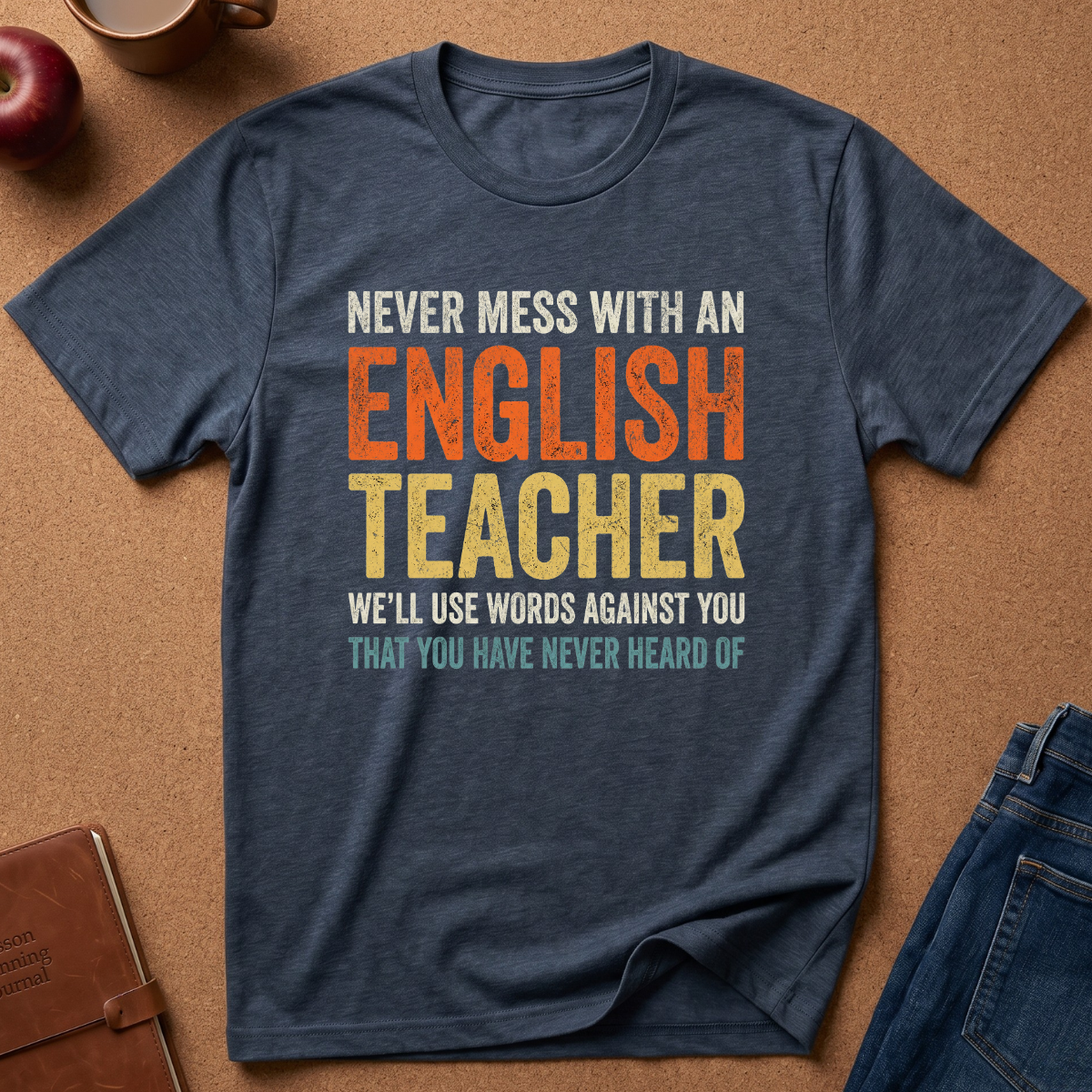 A navy blue graphic t-shirt laid flat on a cork background, featuring a humorous text print that reads 'NEVER MESS WITH AN ENGLISH TEACHER WE'LL USE WORDS AGAINST YOU THAT YOU HAVE NEVER HEARD OF' in distressed orange, yellow, and teal lettering. Styled flat-lay props include a brown leather notebook, a red apple, a coffee mug, and blue jeans.
