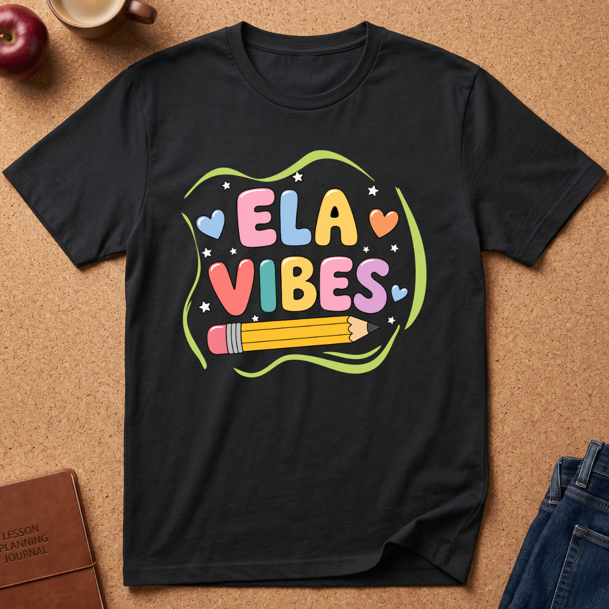 A black short-sleeve graphic T-shirt laid flat on a cork surface, featuring the words 'ELA VIBES' in pastel multicolor letters with a yellow pencil and green wavy outline. Styled flat-lay props include an apple, coffee cup, journal, and jeans around the shirt.