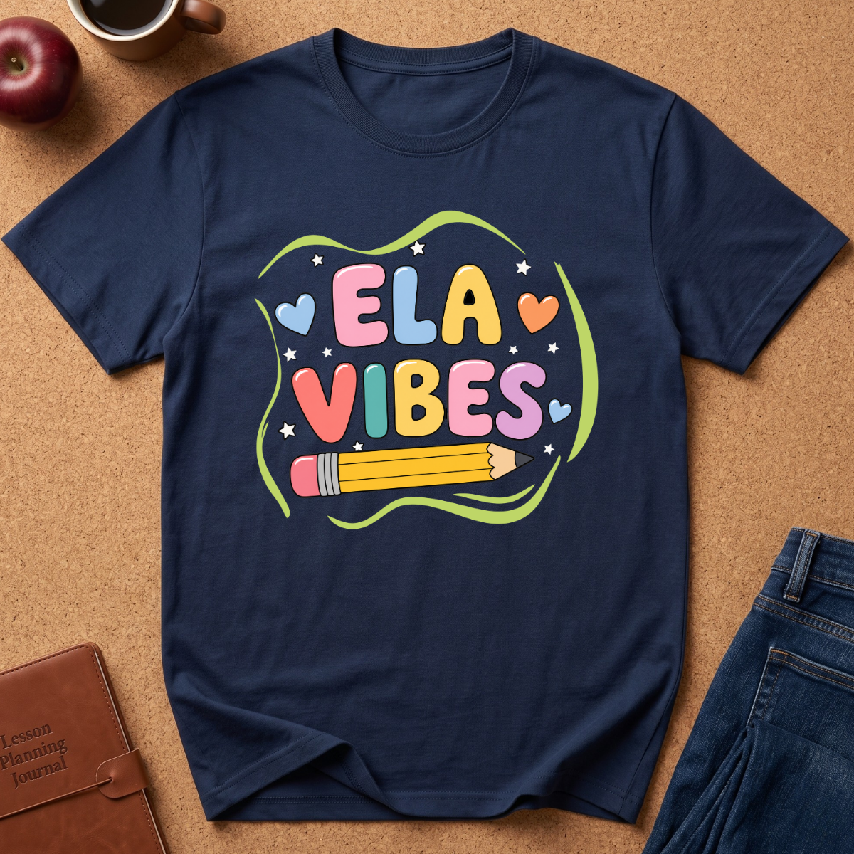 A navy blue graphic t-shirt laid flat on a cork background, featuring a colorful 'ELA VIBES' design with a pencil and hearts. Styled flat-lay props include an apple, a coffee cup, a brown journal, and blue jeans.