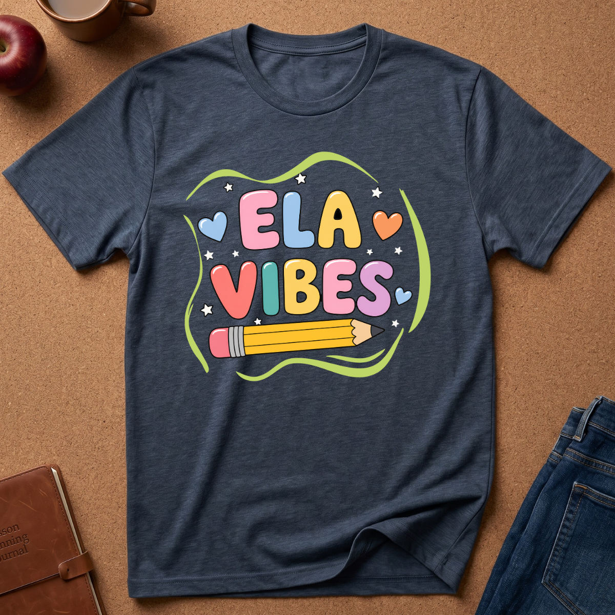 A navy blue heather graphic t-shirt laid flat on a brown cork background, featuring a colorful 'ELA VIBES' school-themed design with a pencil and hearts. Styled flat-lay props include an apple, a coffee mug, a brown notebook, and blue jeans.
