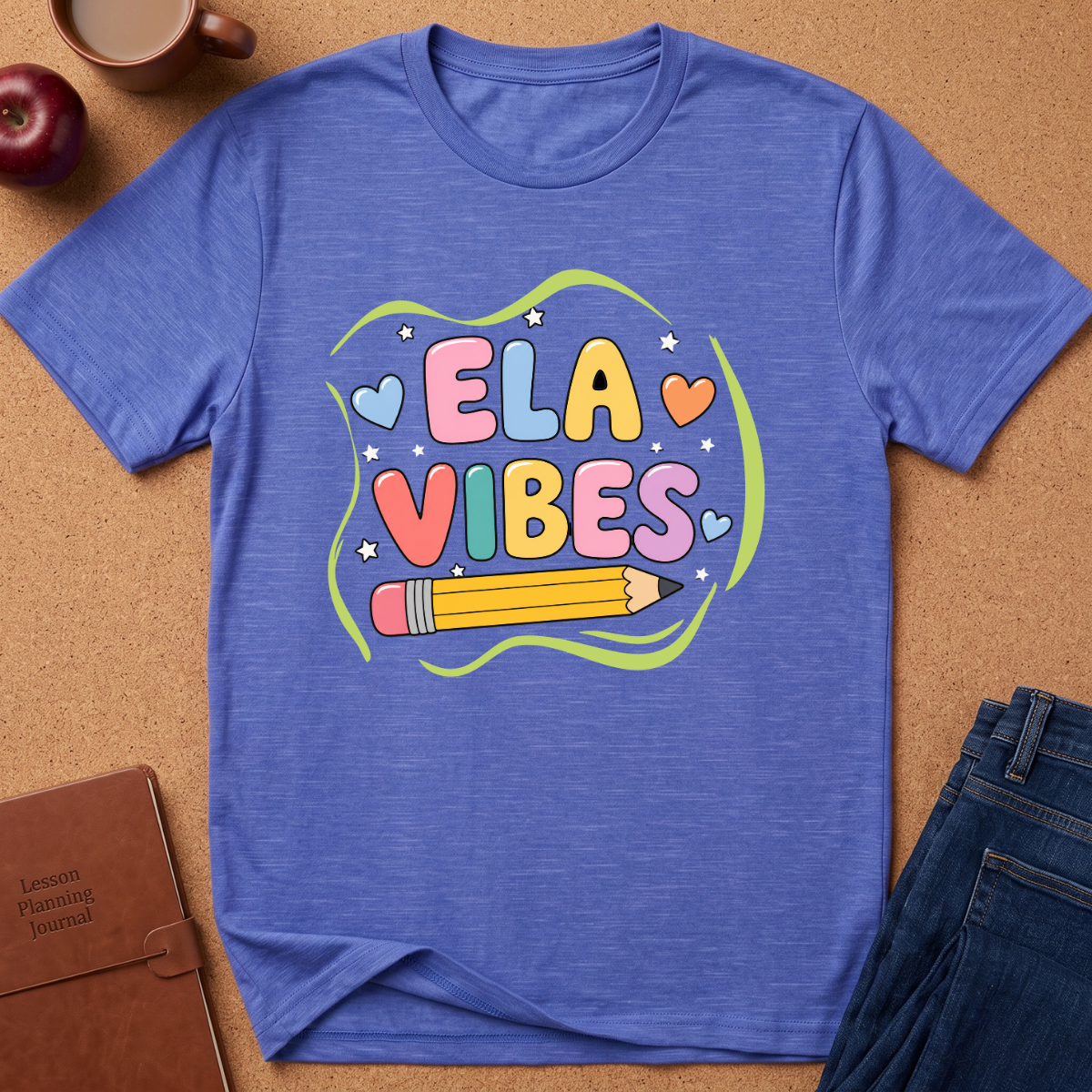 A heathered blue short-sleeve t-shirt laid flat on a cork background, featuring a colorful 'ELA VIBES' graphic with a pencil and hearts. Styled flat-lay props include a brown leather journal, a red apple, a coffee mug, and blue jeans.