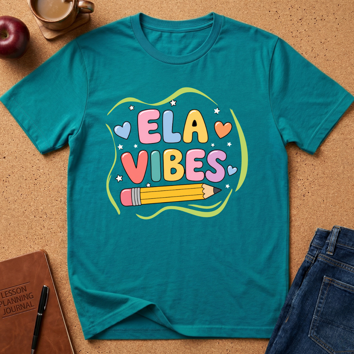 A teal short-sleeve graphic T-shirt laid flat on a cork surface, surrounded by styled props including a coffee cup, apple, notebook, pen, and jeans. The shirt features a colorful 'ELA VIBES' school-themed graphic with hearts, stars, and a pencil.