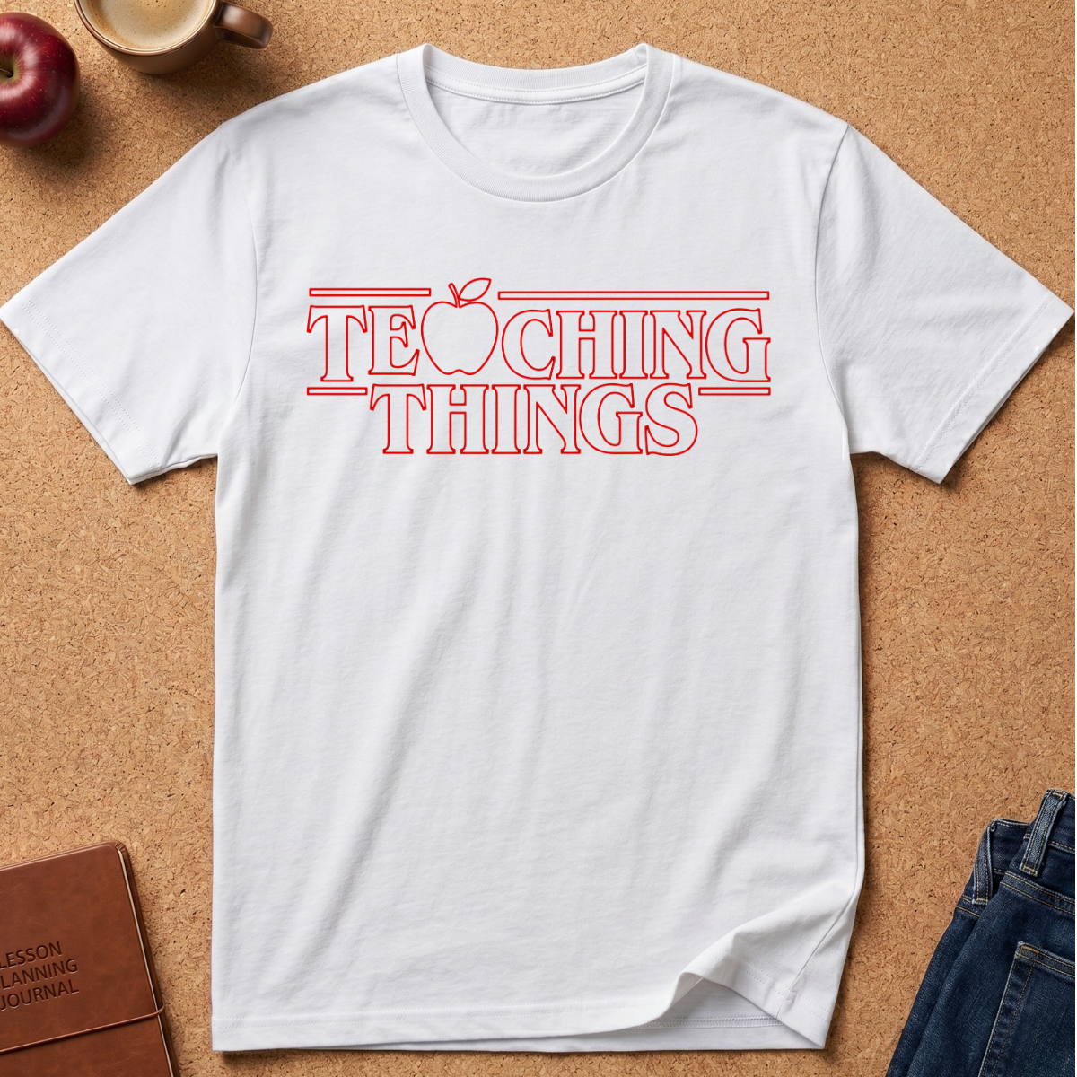 A white t-shirt with a red graphic text reading 'TEACHING THINGS' styled flat-lay on a brown cork background with props like an apple, notebook, and jeans.