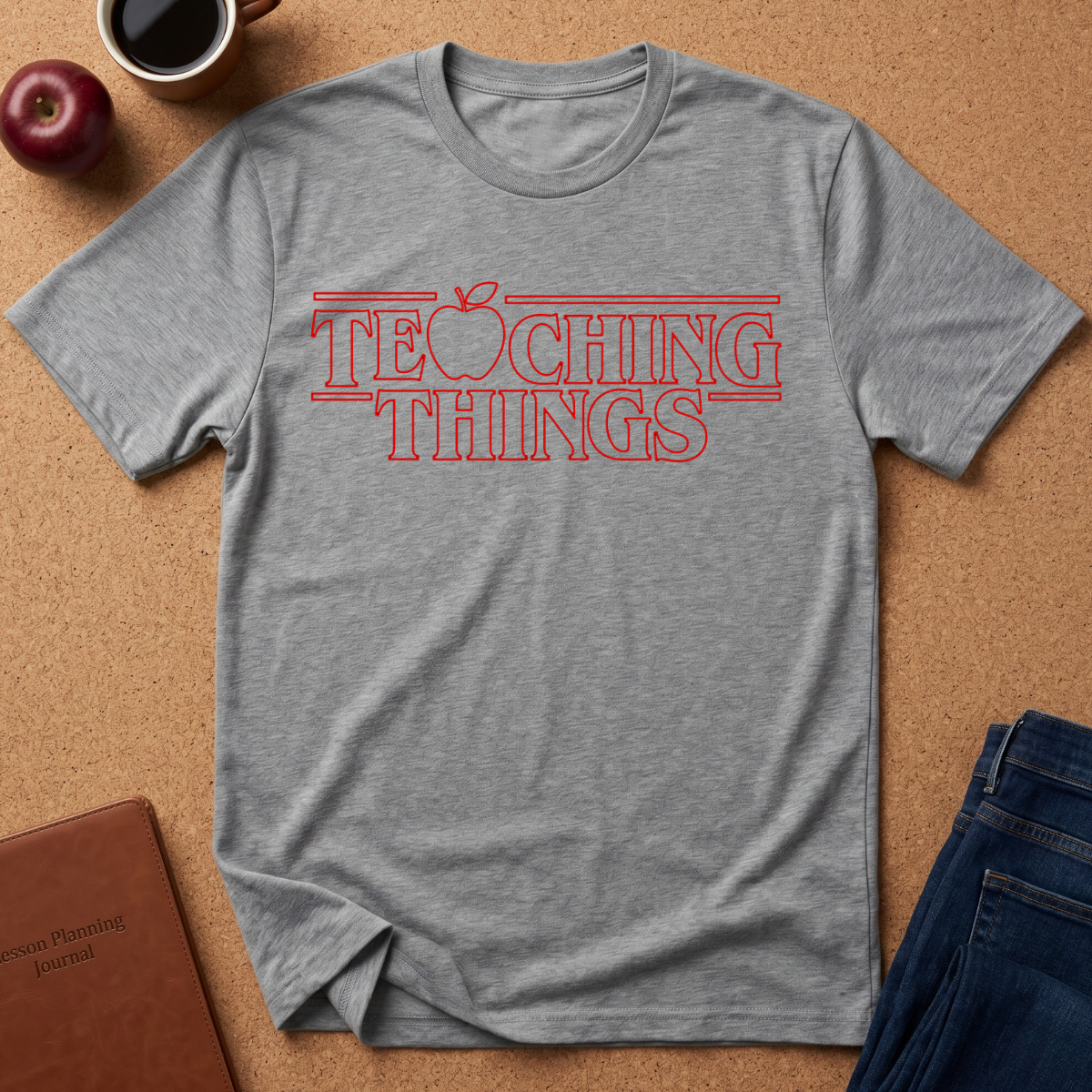 A heathered sports grey t-shirt with a red graphic text reading 'TEACHING THINGS' styled flat-lay on a brown cork background with props like an apple, notebook, and jeans.