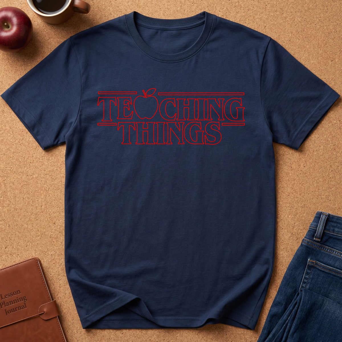 A navy blue t-shirt with a red graphic text reading 'TEACHING THINGS' styled flat-lay on a brown cork background with props like an apple, notebook, and jeans.
