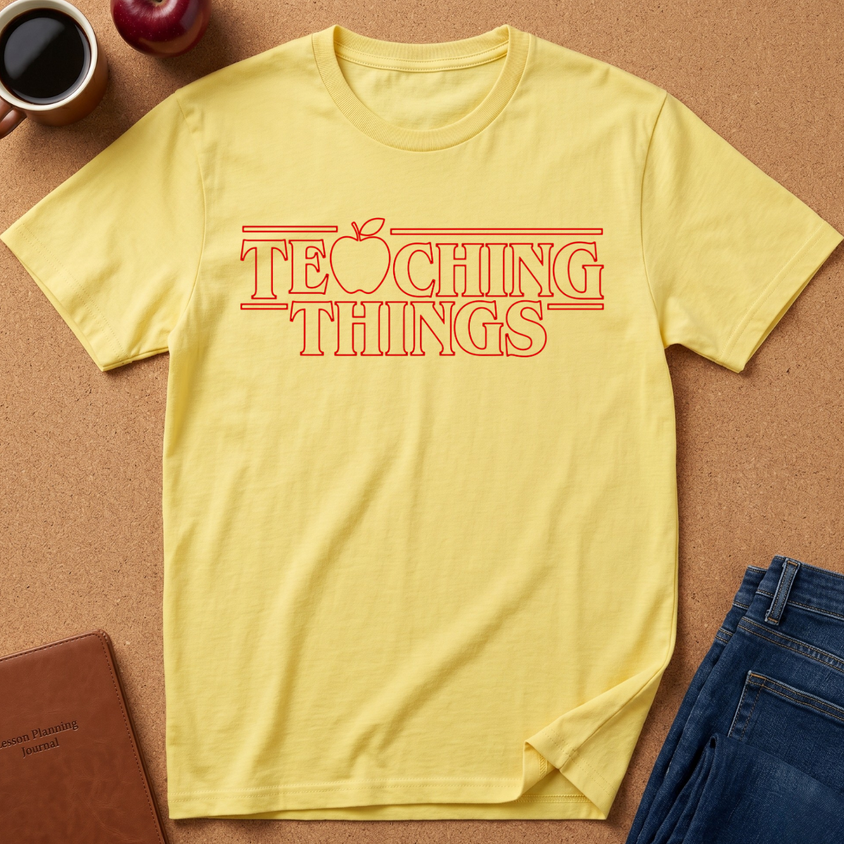 A light yellow  t-shirt with a red graphic text reading 'TEACHING THINGS' styled flat-lay on a brown cork background with props like an apple, notebook, and jeans.
