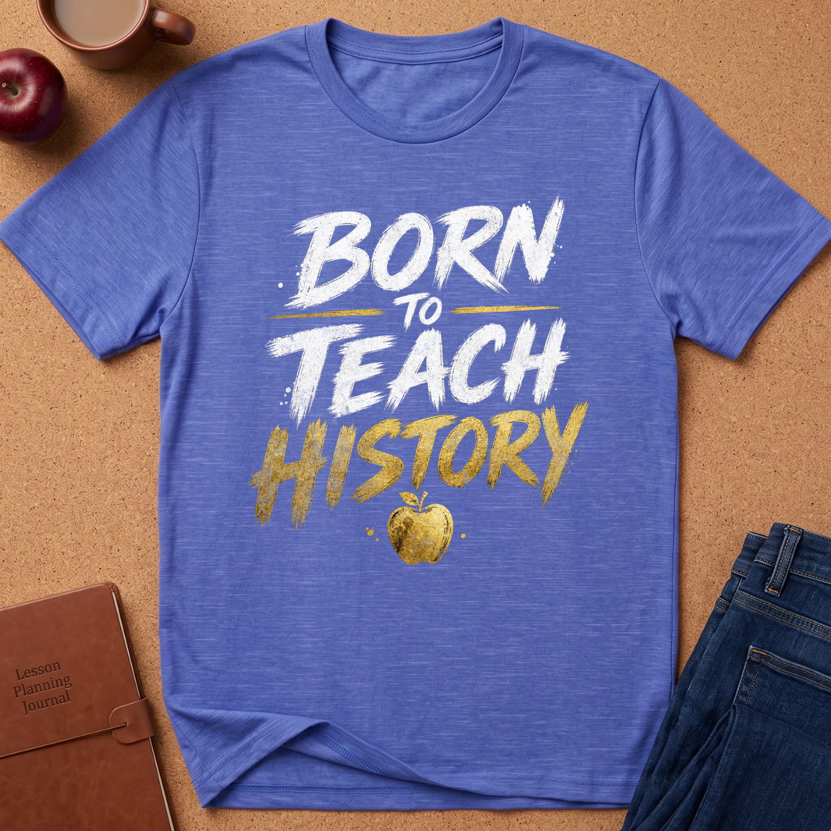 A heathered blue short-sleeve t-shirt with a 'BORN TO TEACH HISTORY' graphic in white and gold, styled in a flat lay on a cork background with an apple, coffee mug, notebook, and jeans.