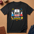 A black short-sleeve graphic T-shirt laid flat on a cork surface, featuring a colorful pop-quizzing-themed design with a periodic-table-style 'Ah!' panel, beakers, stars, and the text 'Discovered during pop quizzes.' Partial props around the shirt include an apple, a mug, a notebook, and jeans.