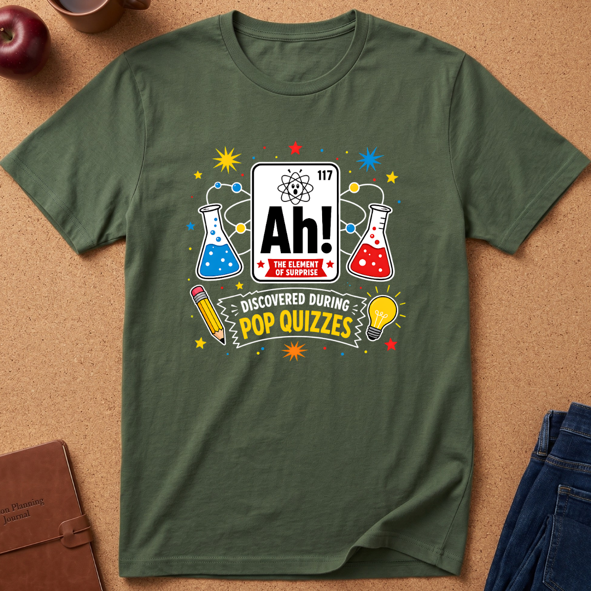 An olive green short-sleeve t-shirt with a colorful science-themed graphic reading 'Ah! The Element of Knowledge' and 'Discovered During Pop Quizzes,' laid flat on a cork background with small props including an apple, notebook, and jeans.