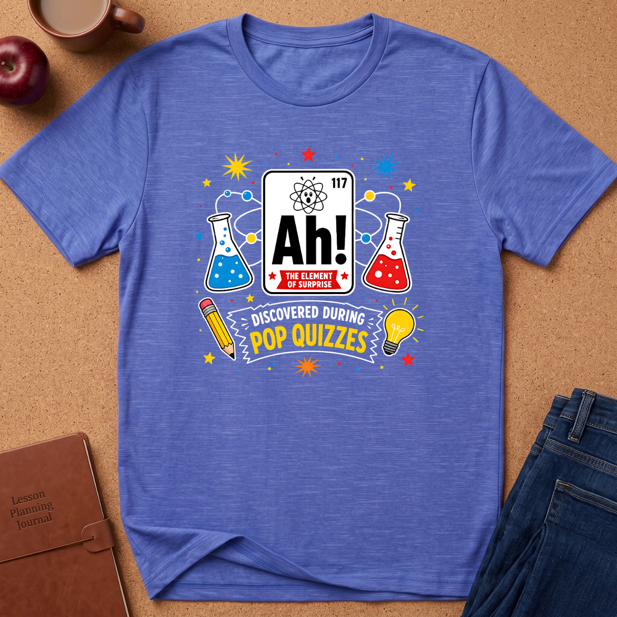 A heathered blue crewneck t-shirt laid flat on a cork background, featuring a colorful 'Ah! The Element of Pop Quizzes' graphic with beakers, a lightbulb, and a pencil.