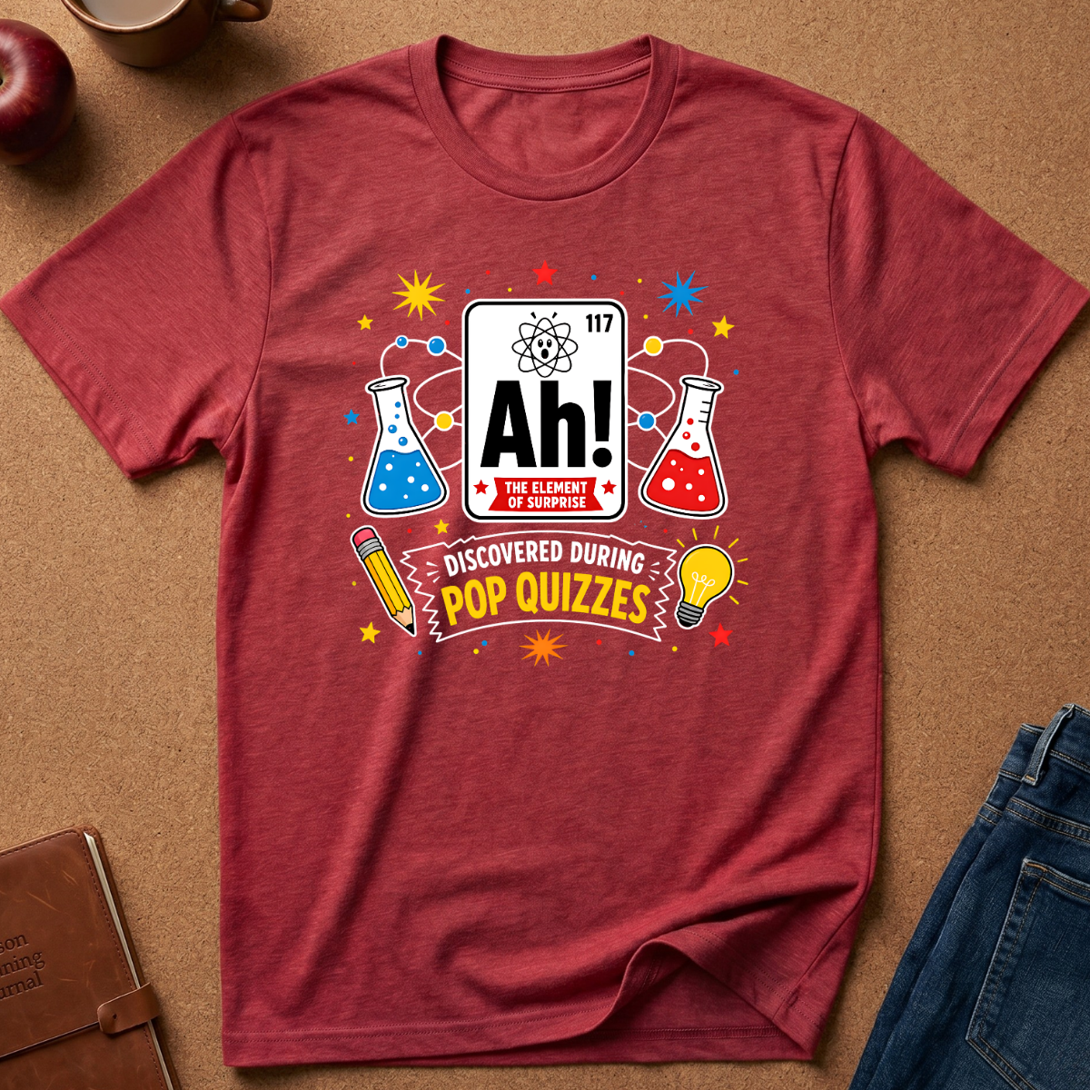 A heathered red short-sleeve t-shirt laid flat on a brown cork-like surface, featuring a colorful pop-quizzes-themed graphic with a periodic-table-style 'Ah!' element, beakers, stars, and the text 'DISCOVERED DURING POP QUIZZES.' Partial jeans, an apple, a mug, and a notebook are visible around the shirt as styled props.