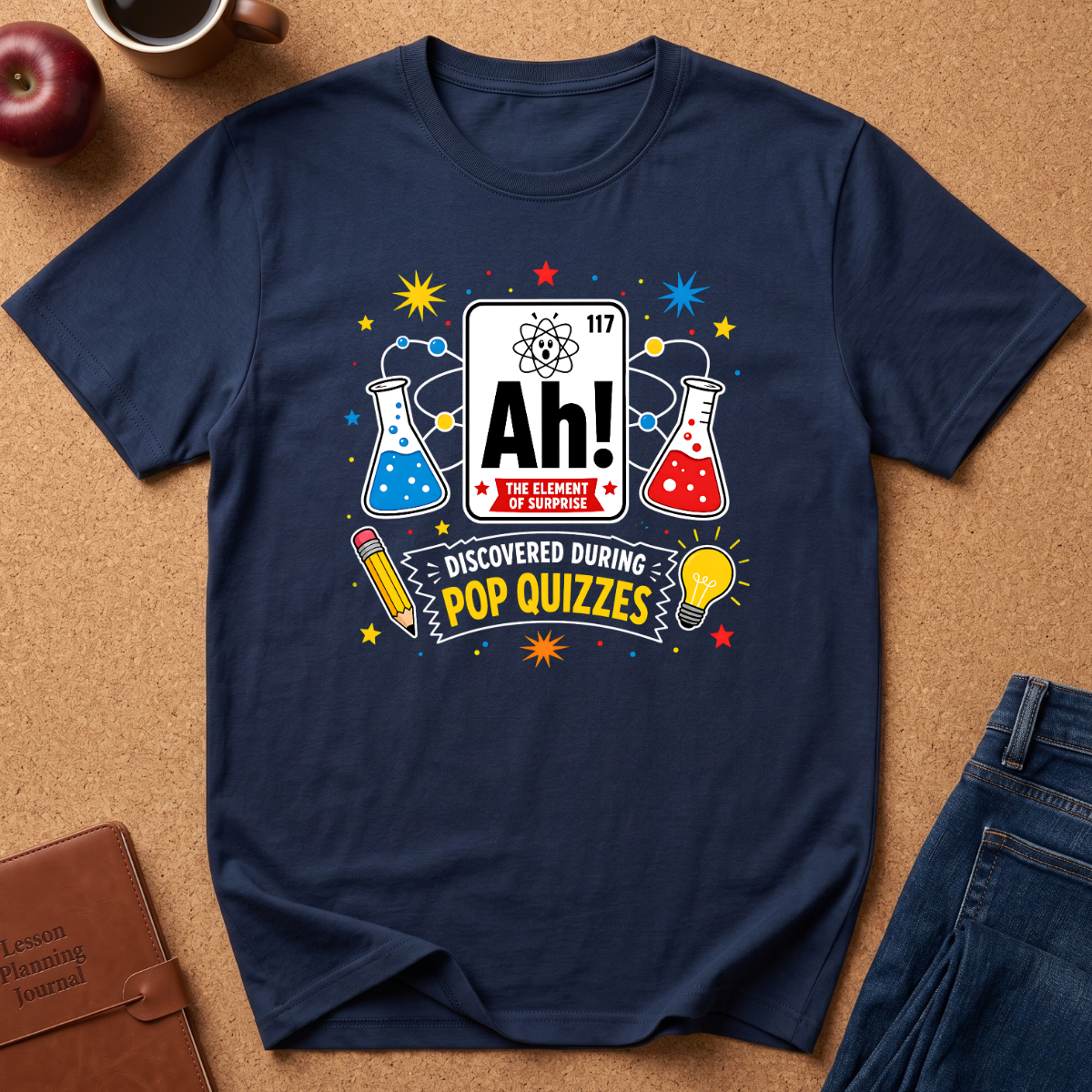 A navy blue short-sleeve t-shirt laid flat on a brown cork background, featuring a colorful science-themed graphic that reads 'Ah! The Element of Knowledge' and 'Discovered during pop quizzes' with beakers, stars, and a lightbulb.