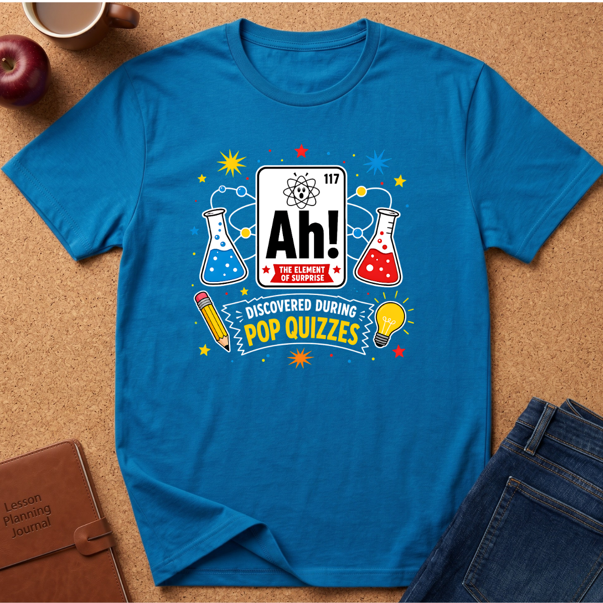 A bright blue short-sleeve graphic T-shirt laid flat on a cork background, surrounded by styled props including an apple, a coffee cup, a notebook, and folded jeans. The shirt features a pop-quizzes themed print with a periodic-table-style panel reading 'Ah! The Element of Knowledge,' beakers, stars, a pencil, and a lightbulb.