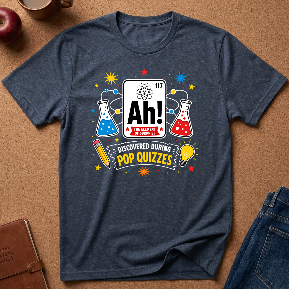 A navy blue graphic t-shirt laid flat on a brown cork background, featuring a colorful 'Ah! The Element of Knowledge' pop culture design with beakers, stars, and a lightbulb.
