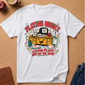 A white short-sleeve graphic T-shirt laid flat on a cork board background, featuring a colorful cartoon desk scene with the text 'PLAYING HOOKY' and 'LESSON PLANS ARE ON THE DESK.' A coffee cup, apple, journal, and jeans are partially visible around the shirt as styled props.