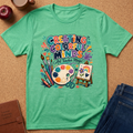 A mint green graphic t-shirt laid flat on a cork background, surrounded by styled props including an apple, a coffee cup, a brown journal, and blue jeans. The t-shirt features a colorful graphic that reads 'CREATING COLORFUL MINDS Art Teacher Magic' with paintbrushes, a paint palette, and an easel.