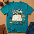 A teal short-sleeve graphic T-shirt laid flat on a cork surface, surrounded by school-themed props including an open notebook with equations, a ruler, pencils, a notebook, and jeans. The shirt graphic reads “SOLVING PROBLEMS DAILY” with math symbols and illustrations.