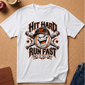 A white short-sleeve graphic T-shirt laid flat on a cork background, featuring a baseball-themed print with crossed bats, a laughing baseball, and the text 'HIT HARD RUN FAST' plus 'Est. 2026'.