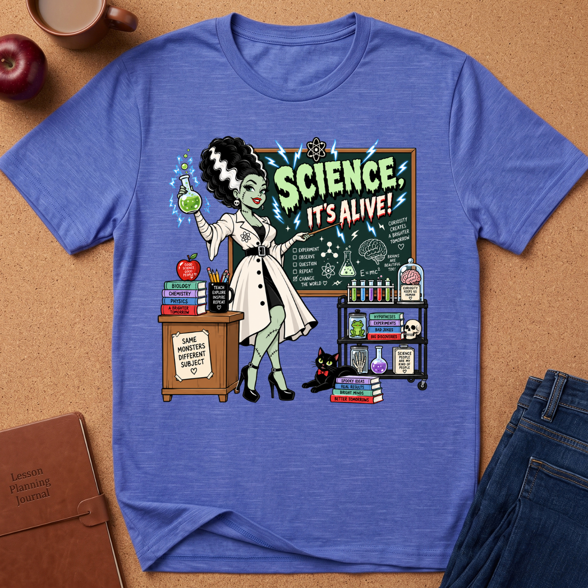 A heathered blue short-sleeve t-shirt laid flat on a brown cork background, featuring a colorful retro graphic of a cartoon scientist in a lab coat with the text 'SCIENCE IT'S ALIVE!'. Surrounding props include an apple, a coffee cup, a notebook, and folded jeans.