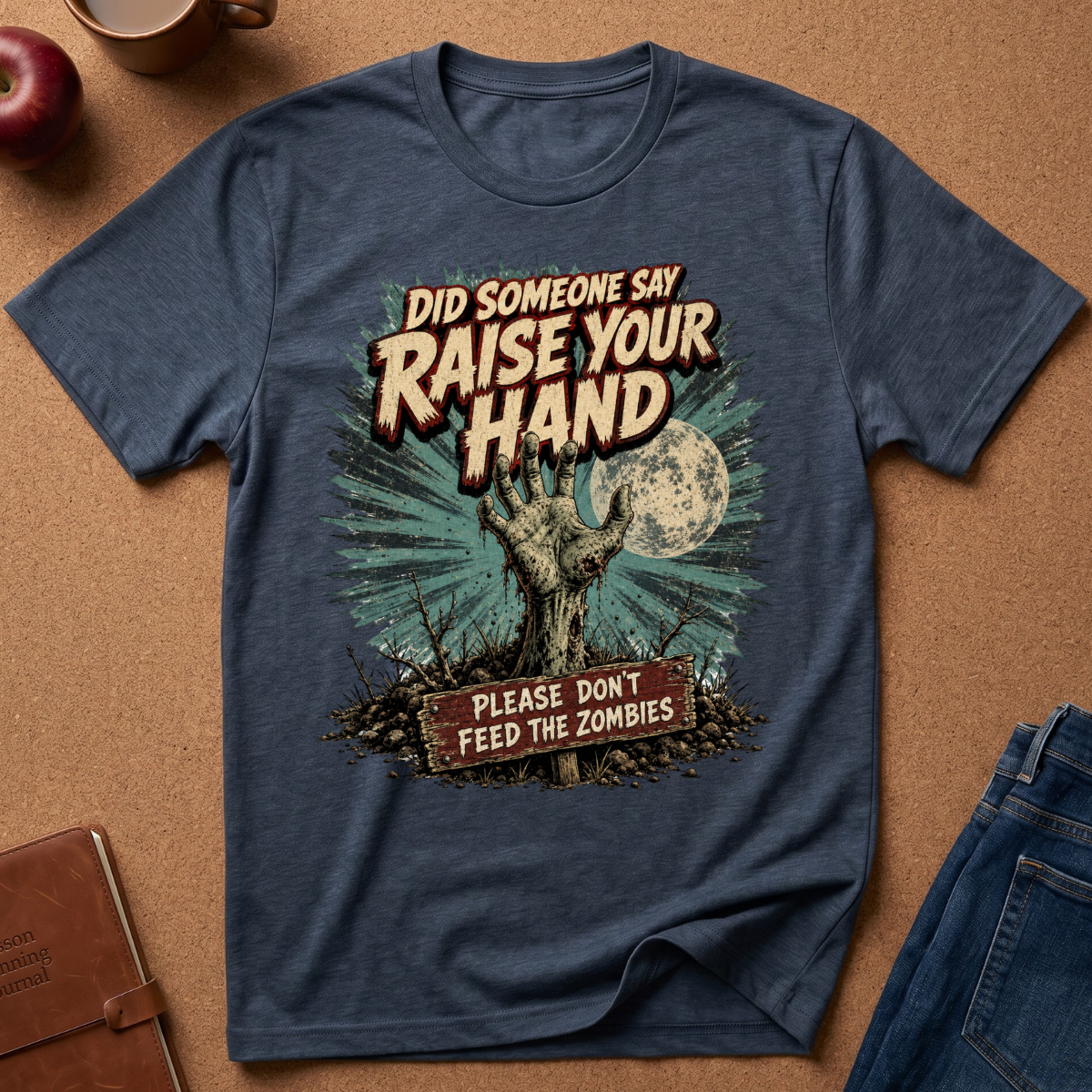 A heathered navy blue graphic t-shirt laid flat on a brown cork background, surrounded by lifestyle props like an apple, a notebook, a coffee mug, and jeans. The t-shirt features a zombie graphic with the text 'DID SOMEONE SAY RAISE YOUR HAND PLEASE DON'T FEED THE ZOMBIES!'.