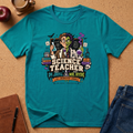 A teal short-sleeve graphic T-shirt laid flat on a cork surface with styled props including an apple, coffee cup, notebook, and jeans. The shirt features a colorful illustrated design of a nerdy science teacher with books, lab equipment, and the text 'SCIENCE TEACHER AND DR.