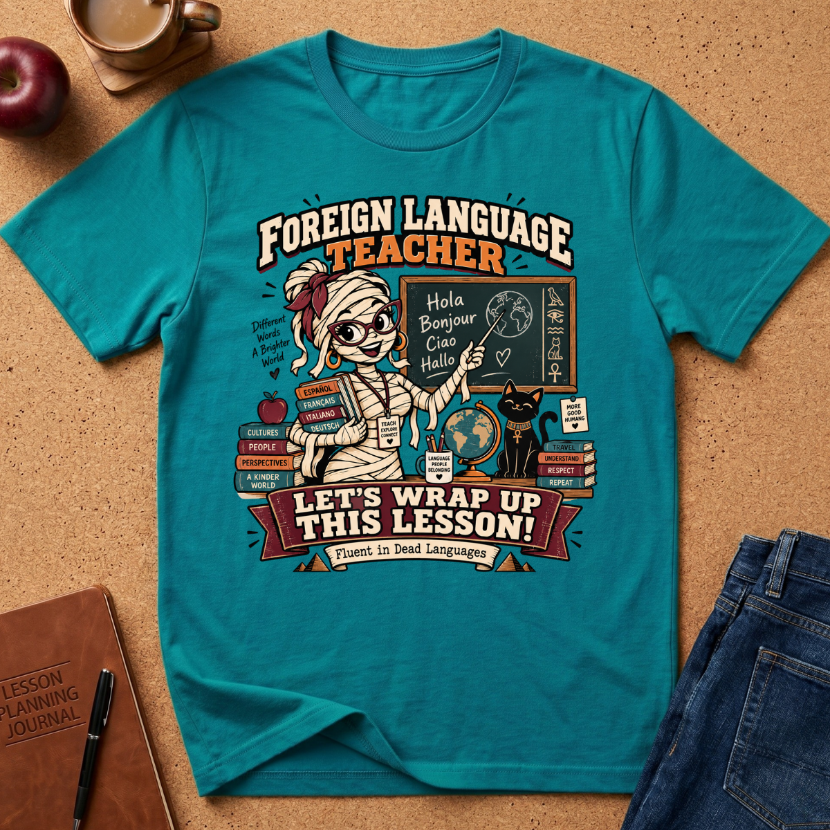 A teal graphic t-shirt laid flat on a cork surface, surrounded by styled props including a notebook, pen, apple, coffee cup, and jeans. The shirt features a cartoon mummy teacher at a chalkboard with the text 'FOREIGN LANGUAGE TEACHER LET'S WRAP UP THIS LESSON!'.