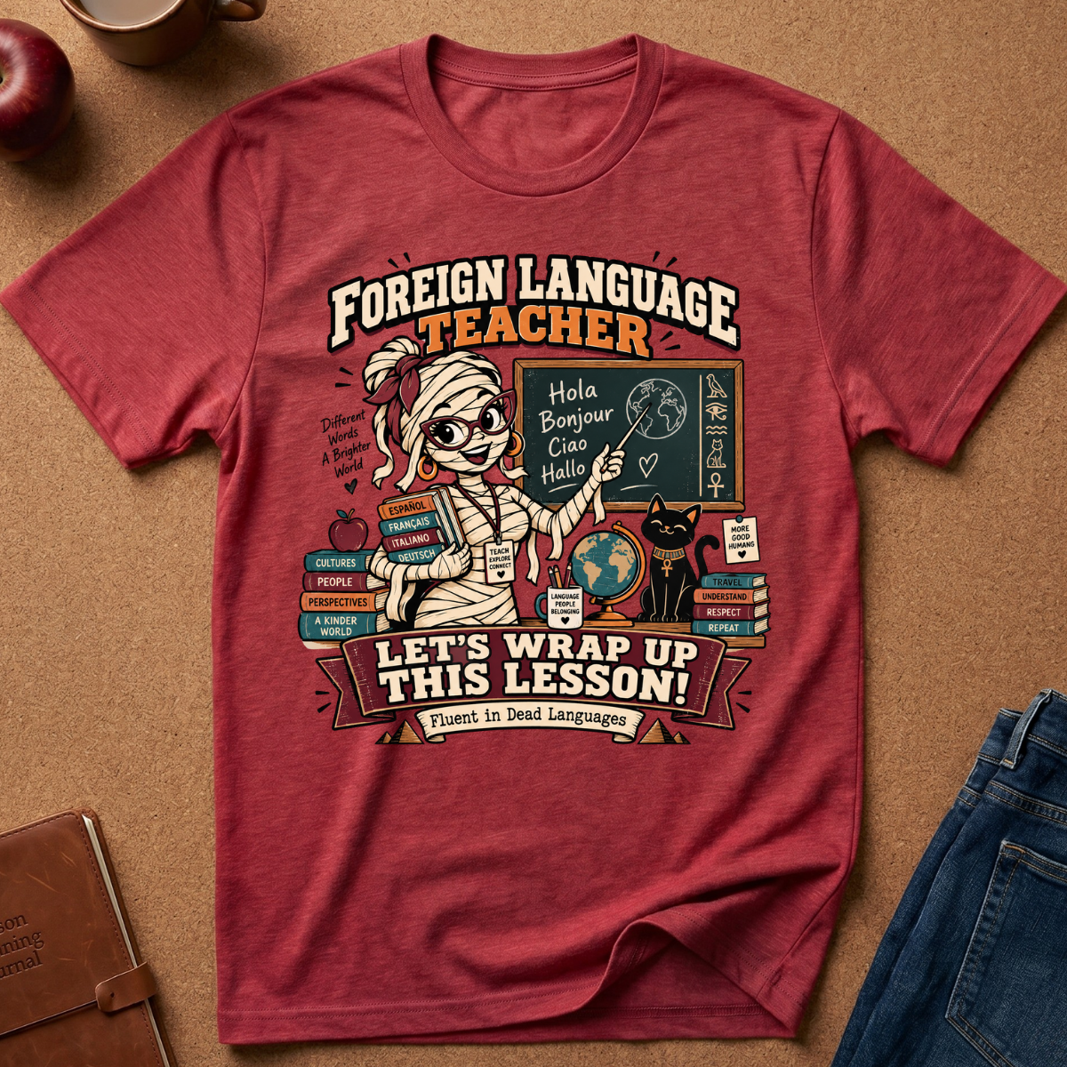 A heathered red graphic t-shirt featuring a cartoon mummy teacher at a chalkboard with the text 'FOREIGN LANGUAGE TEACHER LET'S WRAP UP THIS LESSON! Fluent in Dead Languages', styled in a flat lay on a brown cork background with jeans, a notebook, and an apple.