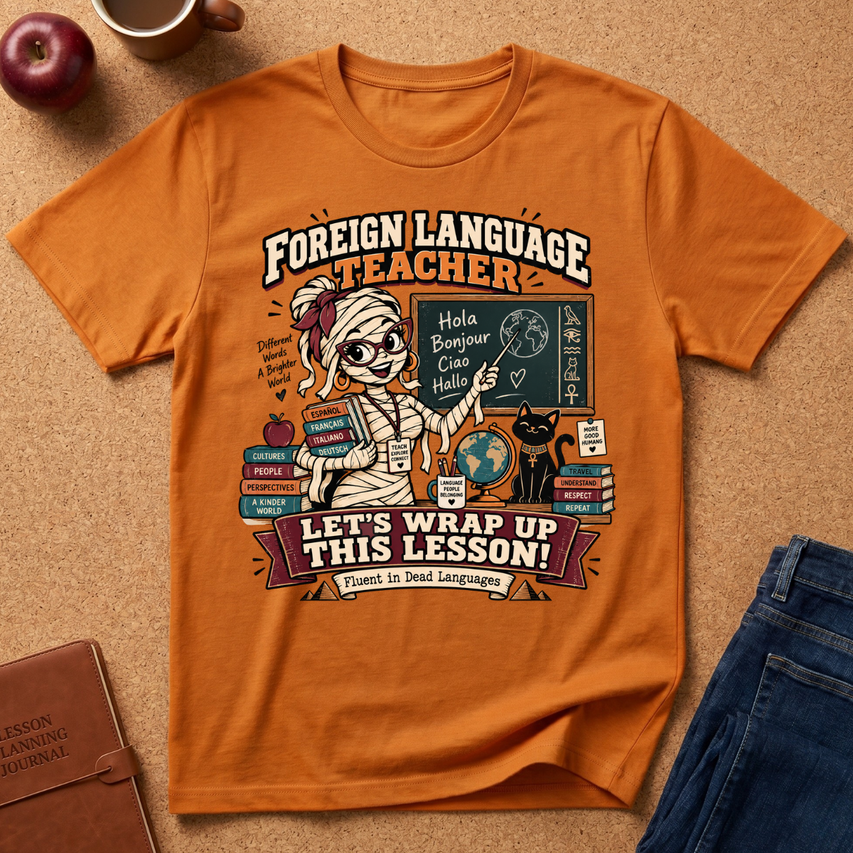 An orange graphic t-shirt featuring a cartoon mummy teacher at a desk with books, chalkboard, and a cat, with the text 'FOREIGN LANGUAGE TEACHER' and 'LET'S WRAP UP THIS LESSON!'. The shirt is styled in a flat lay on a cork background with an apple, notebook, and jeans nearby.