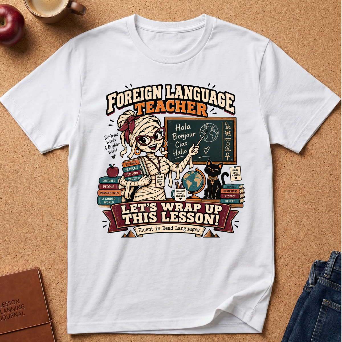 A white graphic t-shirt featuring a cartoon mummy teacher with books, chalkboard, and a cat, with the text 'FOREIGN LANGUAGE TEACHER' and 'LET'S WRAP UP THIS LESSON!'. The shirt is laid flat on a cork background with partial props visible at the edges.