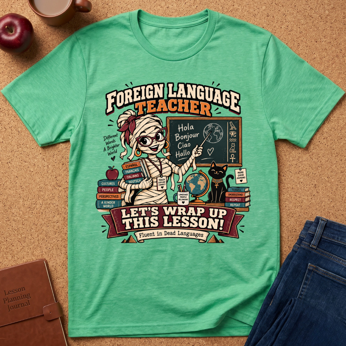 A mint green graphic t-shirt laid flat on a cork background, surrounded by lifestyle props including an apple, a coffee cup, a journal, and blue jeans. The t-shirt features a detailed cartoon graphic of a mummy teacher surrounded by books and a black cat, with the text 'FOREIGN LANGUAGE TEACHER' and 'LET'S WRAP UP THIS LESSON!'.
