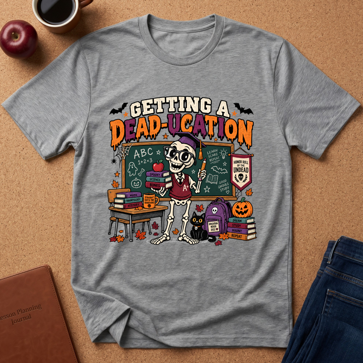 A heather gray graphic t-shirt laid flat on a cork background, featuring a colorful Halloween-themed school scene with the text 'GETTING A DEAD-UCATION'. The design includes a skeleton teacher, chalkboard, books, a black cat, a pumpkin, and bats.
