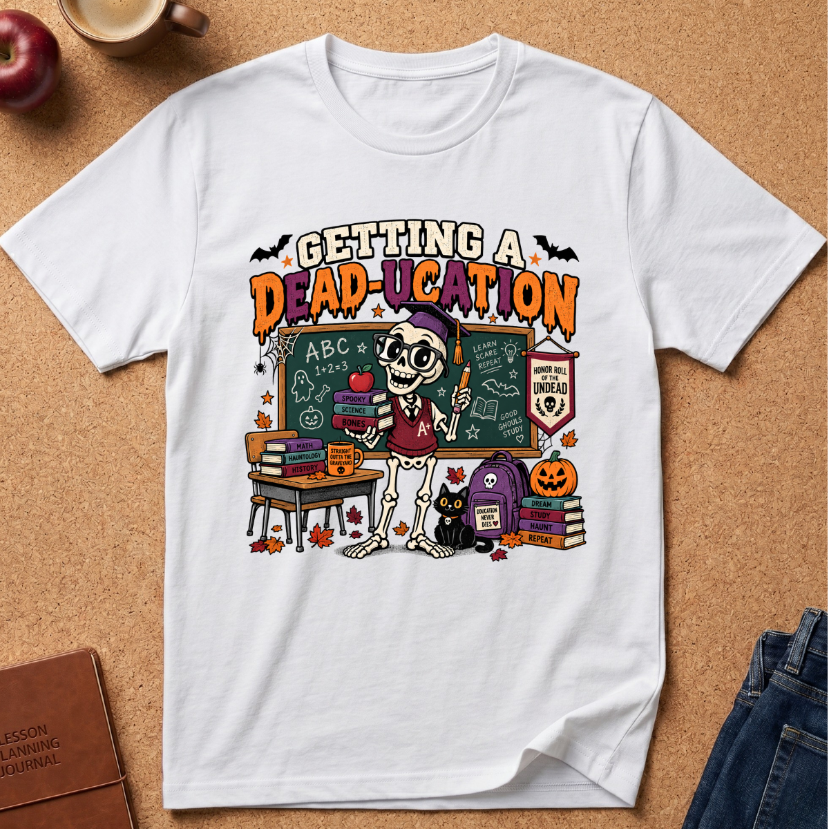 A white short-sleeve graphic T-shirt laid flat on a cork surface, featuring a colorful Halloween-themed school scene with a skeleton teacher, chalkboard, books, pumpkin, and the text 'GETTING A DEAD-UCATION.' Surrounding props include an apple, a coffee cup, a journal, and folded jeans.