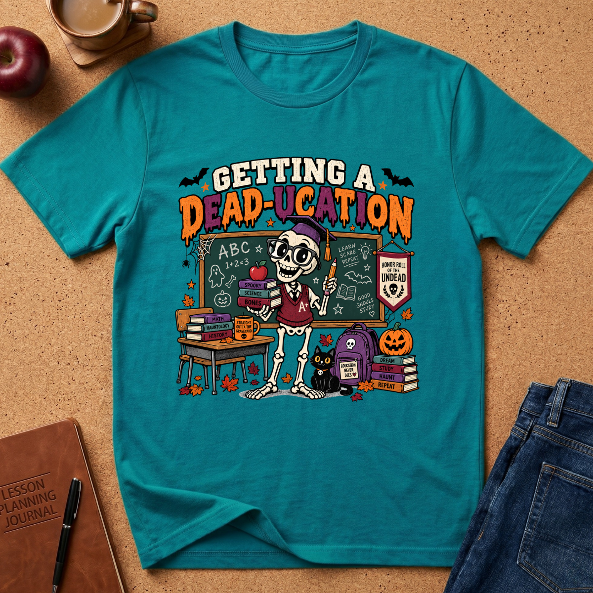 A teal short-sleeve graphic T-shirt laid flat on a cork surface with styled props including an apple, coffee cup, notebook, and jeans. The shirt features a large Halloween-themed school scene print with the text 'GETTING A DEAD-UCATION' and an illustrated skeleton teacher, chalkboard, books, pumpkin, and cat.